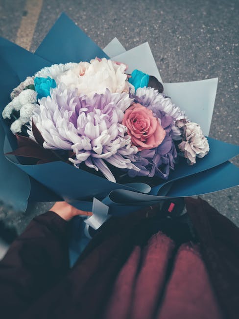 A hand holding a bouquet of fresh flowers wrapped in blue and white paper, featuring a variety of blooms including light purple chrysanthemums, a pink rose, white and blue accents, and green foliage.