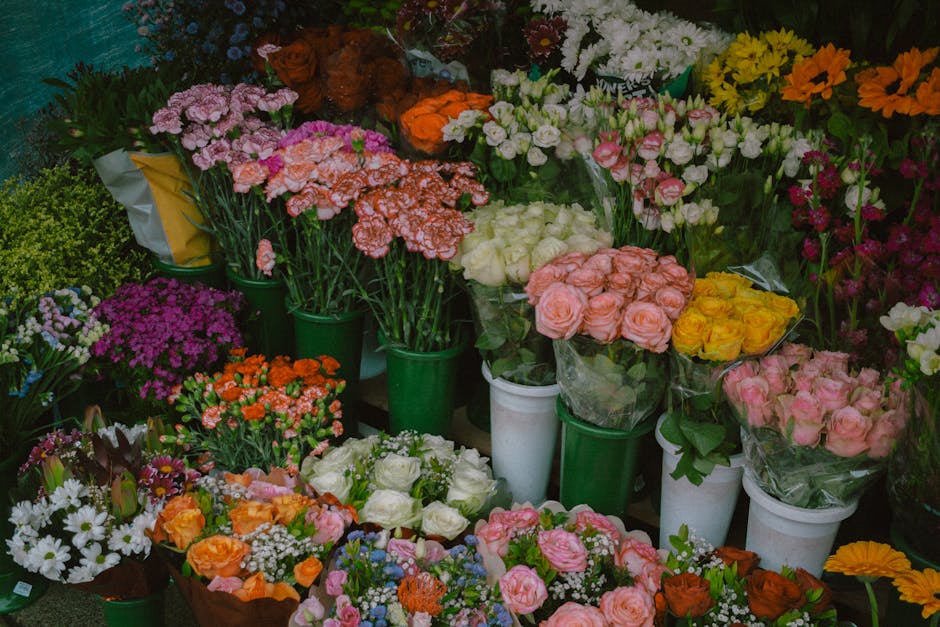 A vibrant display of fresh flowers arranged in various bouquets and individual stems, including pink, yellow, orange, white, and purple blooms. The flowers feature roses, carnations, daisies, and othe
