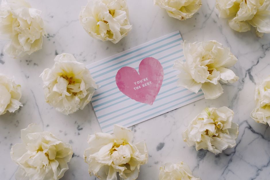 A flat lay of white lisianthus flowers arranged on a white marble surface, surrounding a rectangular card with blue and white stripes and a pink watercolor heart that says 'You're the best' in white t