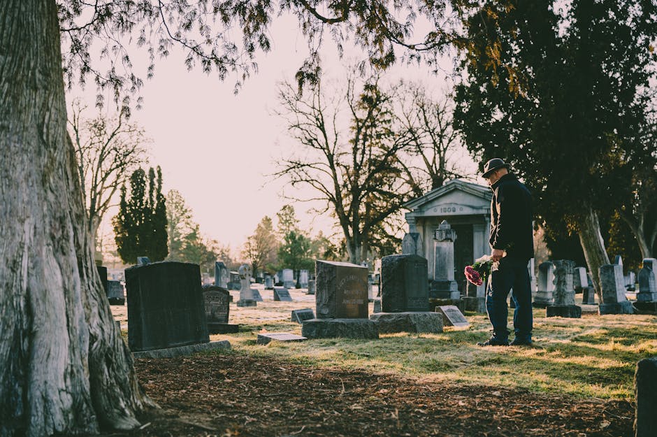 A man dressed in dark formal clothing, including a black hat, stands in a cemetery near Worcester Park station, holding a bouquet of pink and red flowers. The arrangement features fresh, vibrant petal