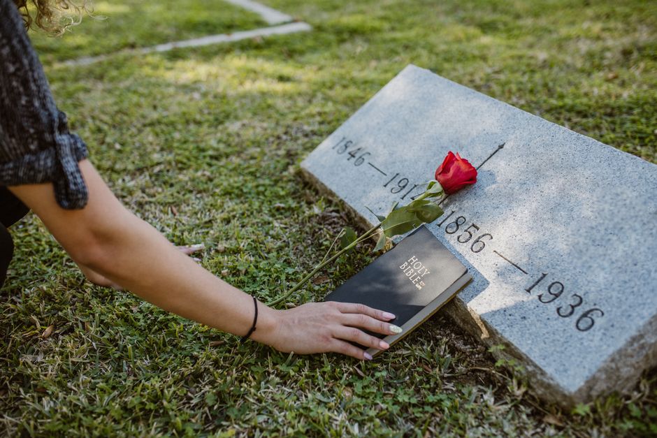 A person placing a fresh, red rose with green leaves on a grey granite memorial stone inscribed with birth and death years, near Worcester Park station, during daylight hours. The person is holding a