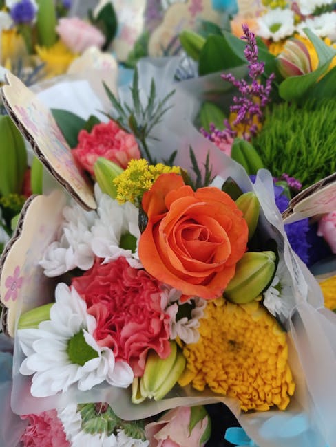 A vibrant floral arrangement featuring a central peach-orange rose with delicate, tightly packed petals, surrounded by white daisies with green centers, bright pink carnations with ruffled petals, yel