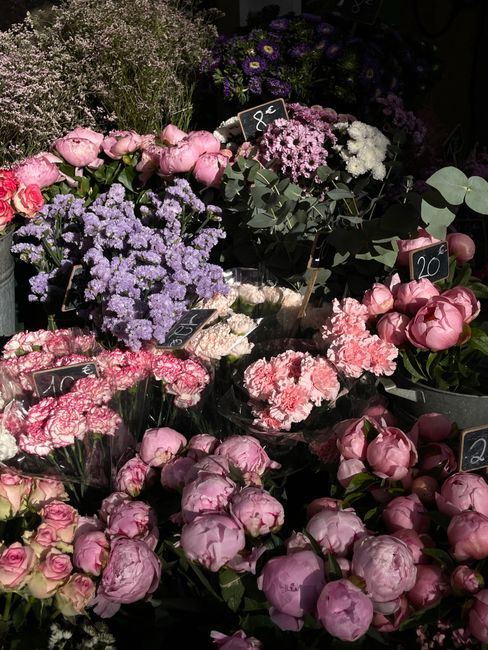 A vibrant display of fresh flowers arranged in various bouquets and containers. The arrangement features clusters of pink peonies with lush, rounded petals, alongside purple and lavender-flowered bloo